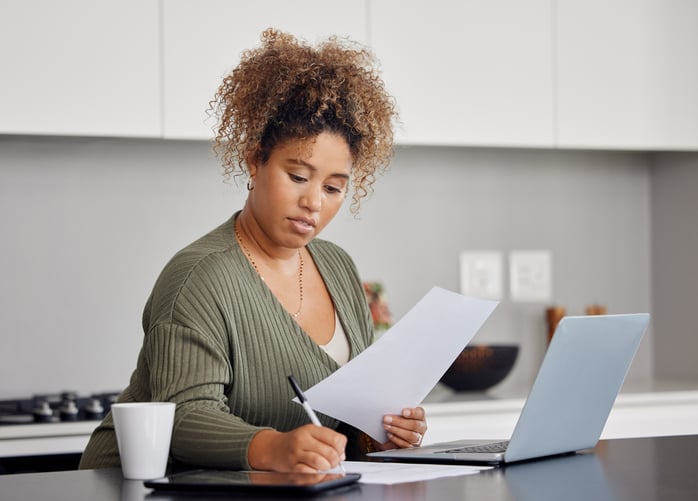 A woman fills out her passport application at home with a pen, paper, and laptop.